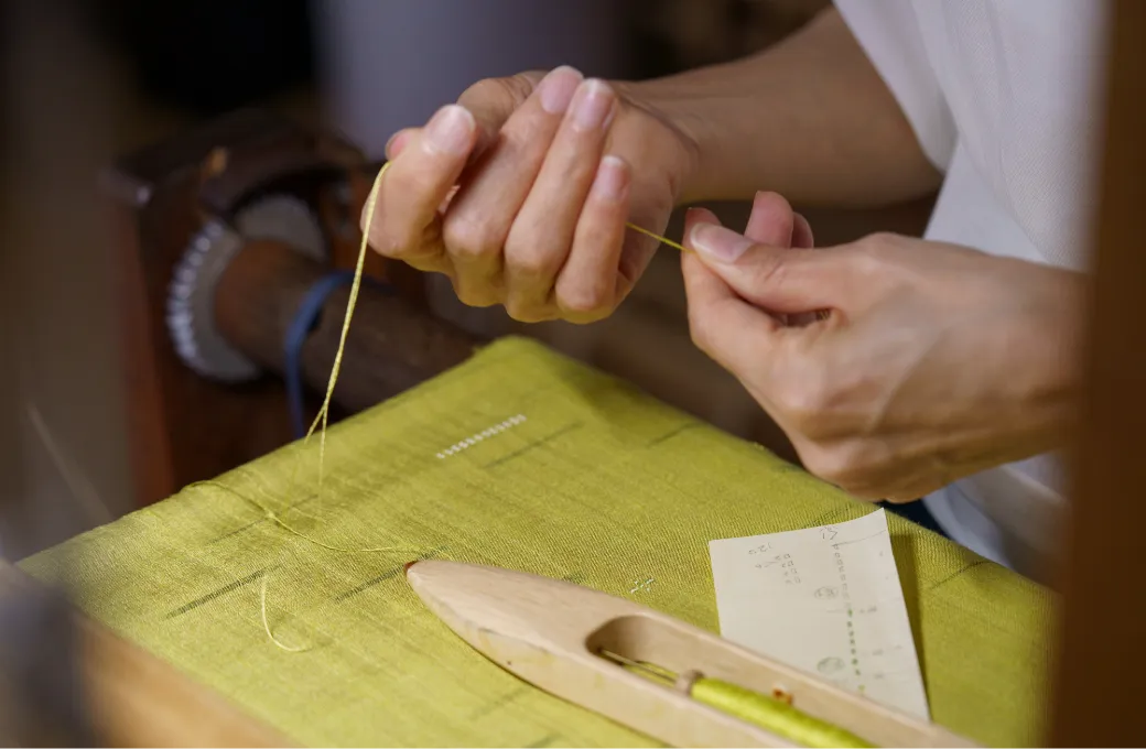 黄緑色の布の上で職人が細い糸を指先で引き揃えている手元の写真。織りや縫製の下準備となる繊細な手仕事の工程が伝わる。
