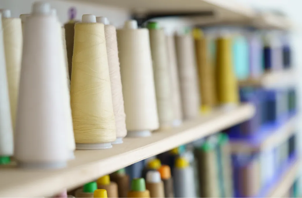色とりどりの工業用ミシン糸のコーンが棚に整然と並んでいる様子。前景は淡い色の糸で、奥に行くほどカラフルな糸が見える。