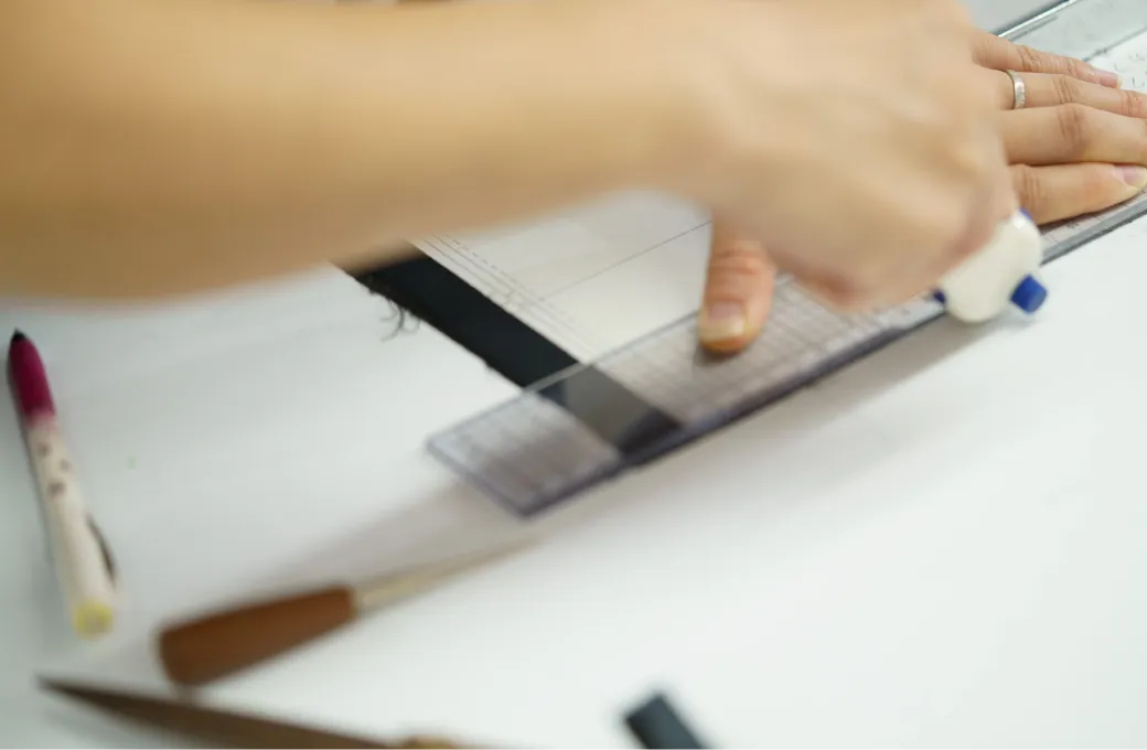 製図用紙の上で定規とペンを使いながら布の上に線を引いている手元。周囲にはチャコペンや裁縫道具が見える。