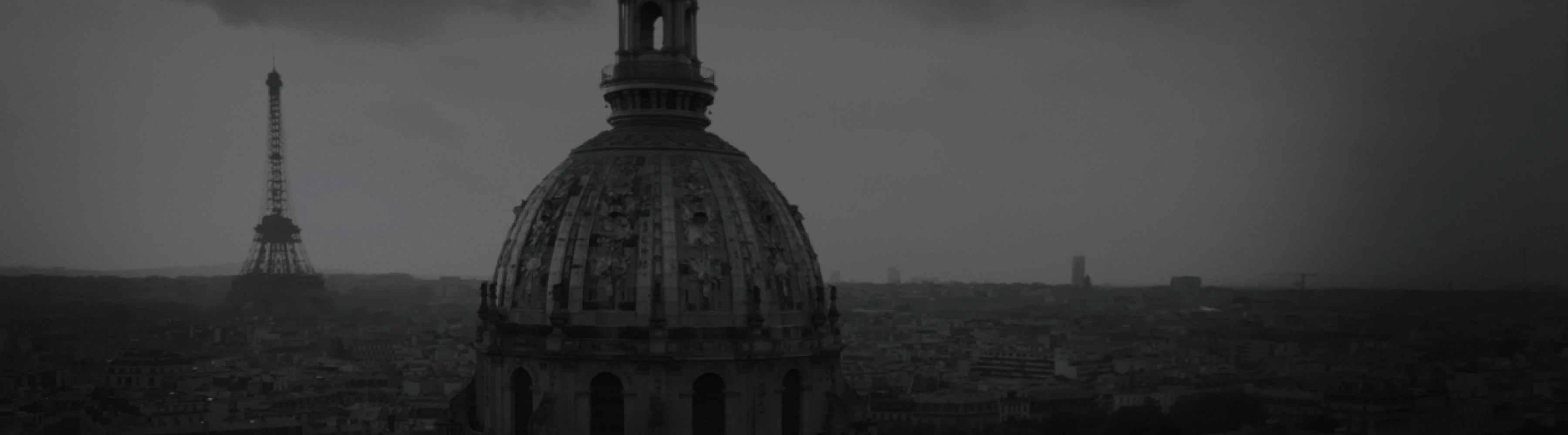 曇り空の下、パリの街並みにそびえるドーム屋根の建物を前景に、左奥にはエッフェル塔がかすんで見えるモノクロ写真。