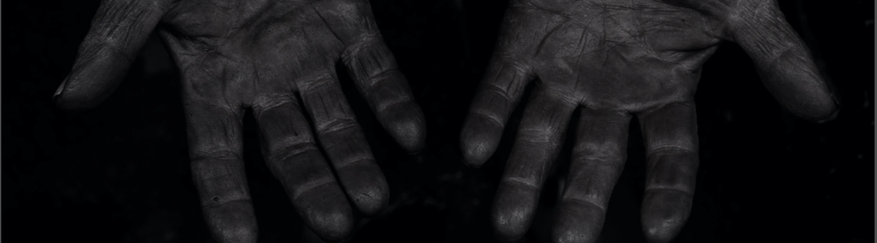 黒く染料が染み込んだように深く汚れた両手のひらを広げて見せているモノクロ写真。しわや質感が強調され、長年の手仕事の跡が感じられる。