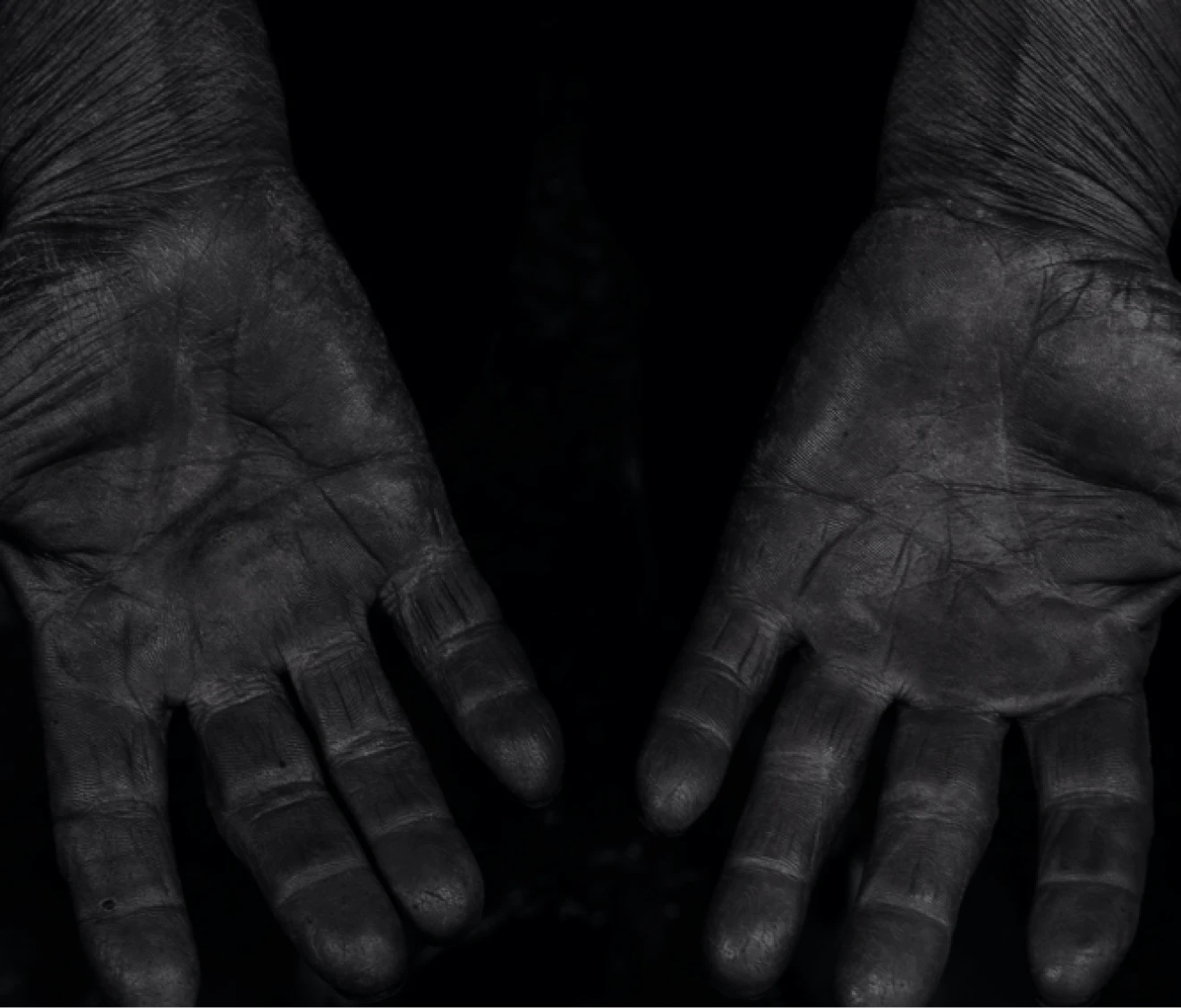 黒く染料が染み込んだように深く汚れた両手のひらを広げて見せているモノクロ写真。しわや質感が強調され、長年の手仕事の跡が感じられる。