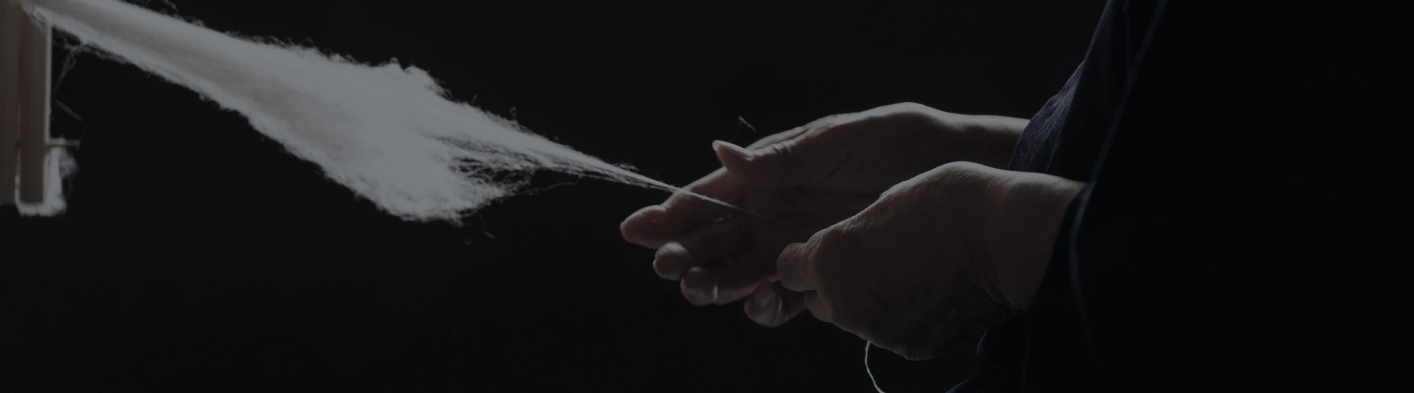 暗い背景の中、職人の手が白い繊維を細く引き出しながら糸にしていく様子を捉えた写真。繊細な指の動きと柔らかな繊維の質感が際立っている。