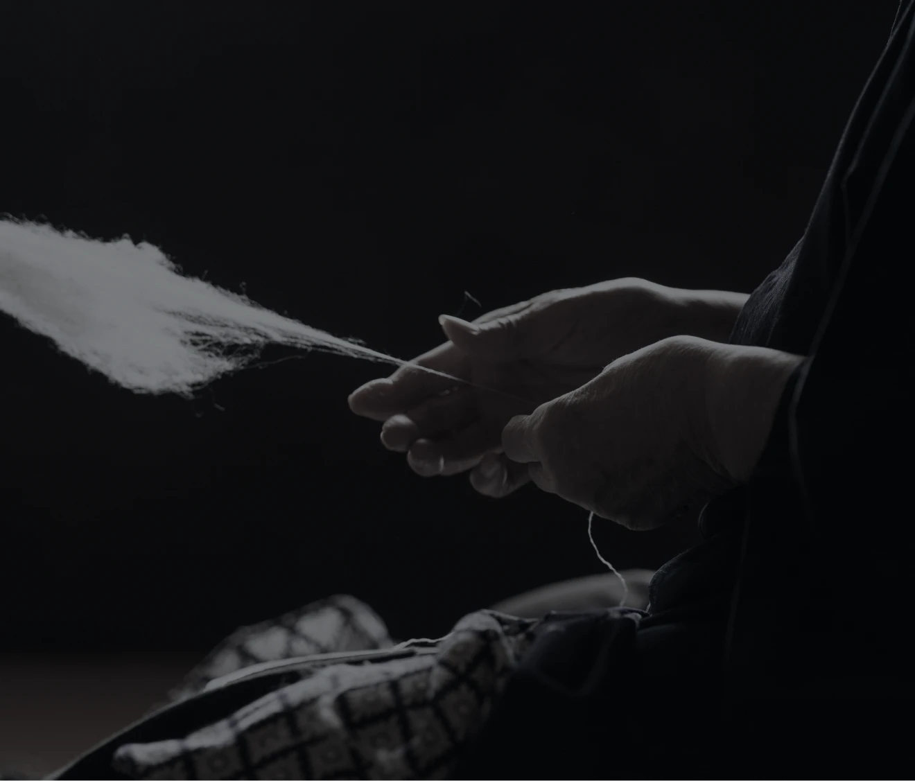 暗い背景の中、職人の手が白い繊維を細く引き出しながら糸にしていく様子を捉えた写真。繊細な指の動きと柔らかな繊維の質感が際立っている。