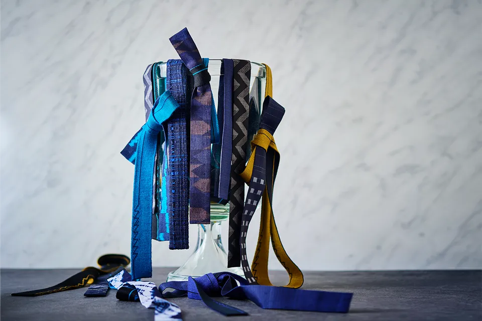 ガラス瓶に藍色や紺、黄色など多彩な柄の細身のネクタイがいくつも掛けられ、幾何学模様や絞り風など異なるデザインが揺れ下がっている。手前のテーブルにも同じ素材のネクタイが広がり、和柄を活かした個性的なネクタイのバリエーションが強調されている。