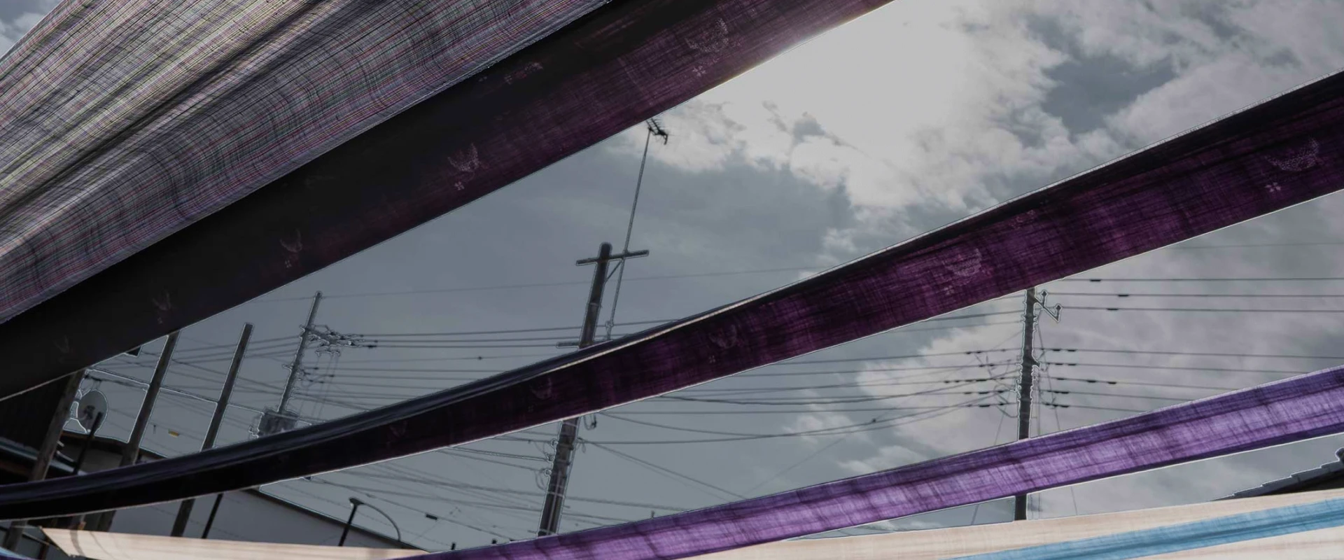 屋外で紫や青などの細長い染め布が空に向かって張られ、その隙間から曇り空と電線、電柱が見える写真。布の透け感と光が重なり合い、伝統的な染色作業の雰囲気が漂っている。