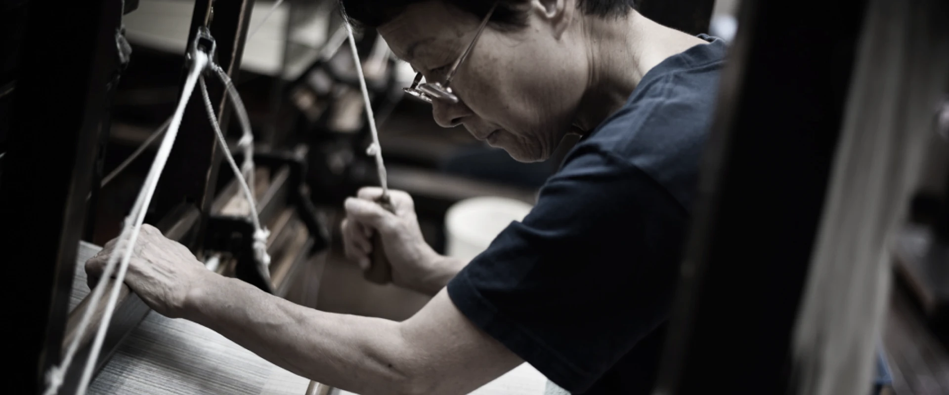 年季の入った織り機に向かい、職人が糸を張りながら真剣な表情で作業している様子。腕や指先の動きから細やかな手仕事の集中が伝わる。