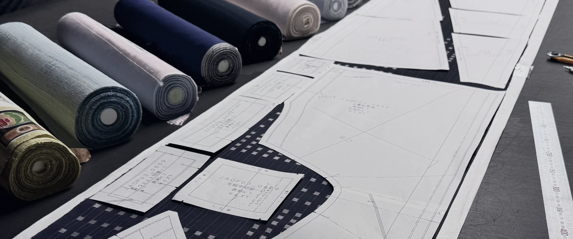 淡い色から濃紺まで並べられた反物の横に、衣服づくりのための型紙が整然と広げられている作業台の写真。丁寧な裁断前の準備工程が伝わる。