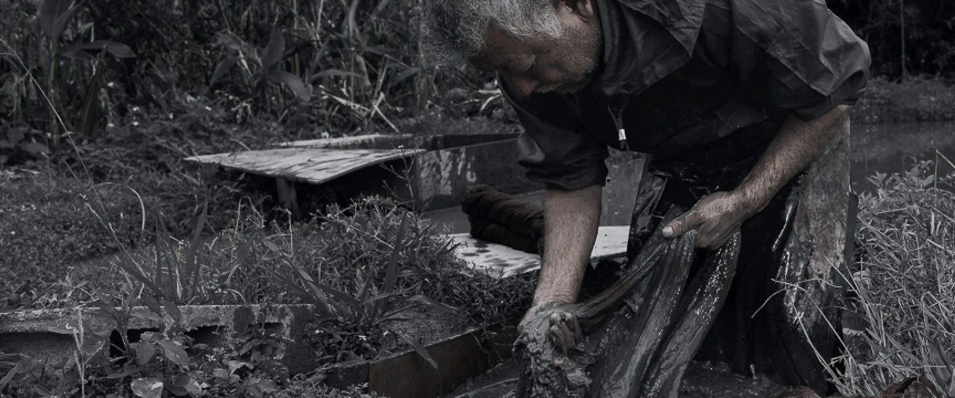 草木に囲まれた屋外で、職人が泥に浸した布を両手で絞りながら染め作業を行っている様子。衣服や手は泥で汚れ、自然の中で行われる伝統的な手仕事の力強さが伝わる。
