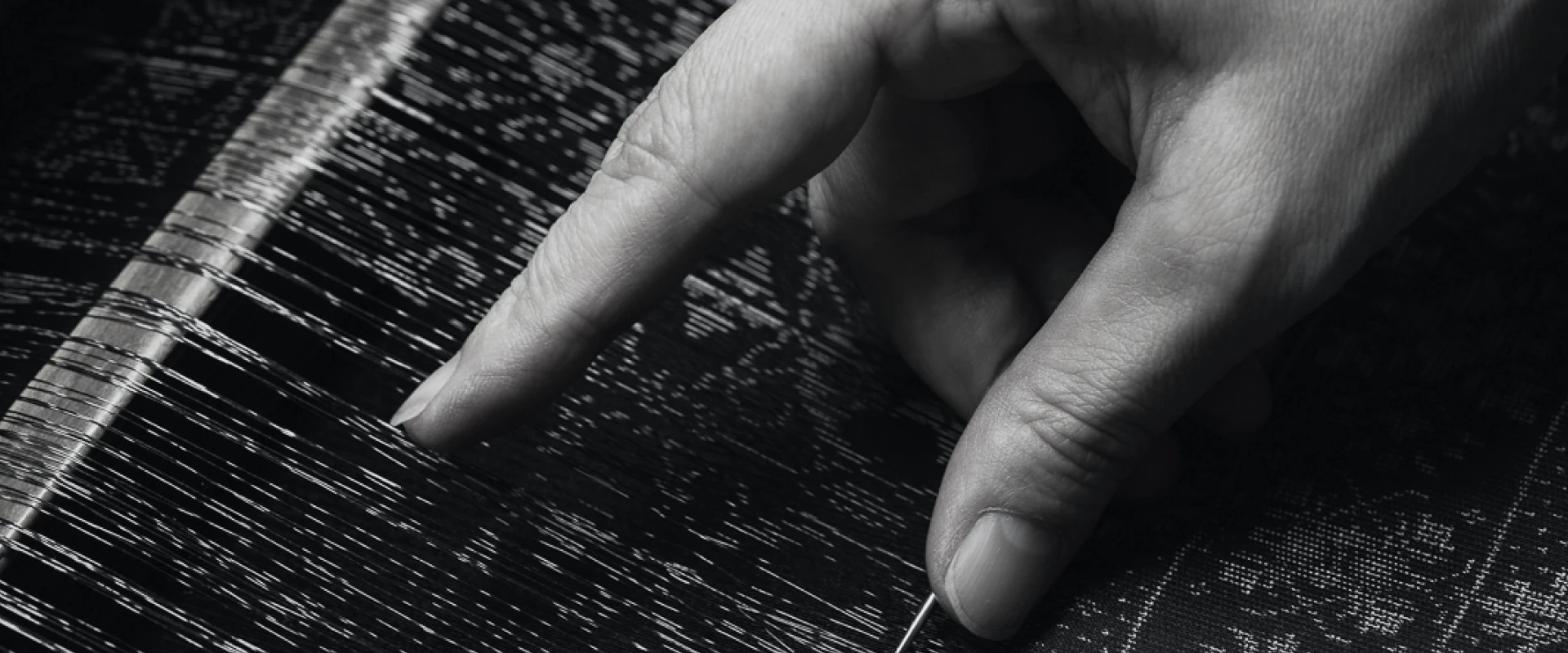 細い糸が張られた織り機の上で、職人の指が一本一本の糸の位置を丁寧に整えているモノクロの手元写真。糸の緊張感と繊細な作業が強調されている。
