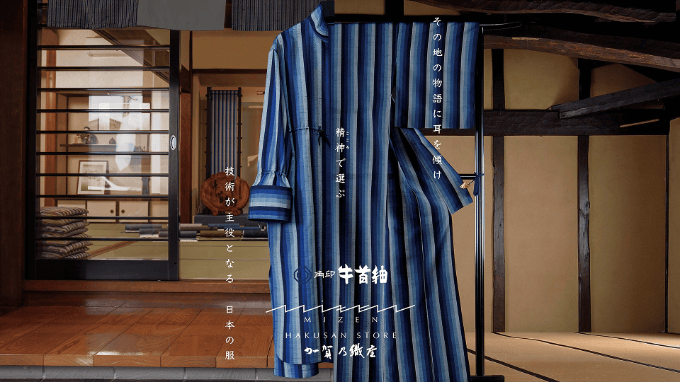伝統的な和室の前に、青と紺の縞模様が美しい牛首紬の反物と、それで仕立てた衣が掛けられている。背景には木枠の引き戸越しに反物や道具が並び、「その地の物語に耳を傾け 精神で選ぶ 技術が主役となる 日本の服」「牛首紬 × MIZEN 白山店 加賀乃織座」などの文字が配置され、職人技と地域文化を伝えるビジュアルになっている。
