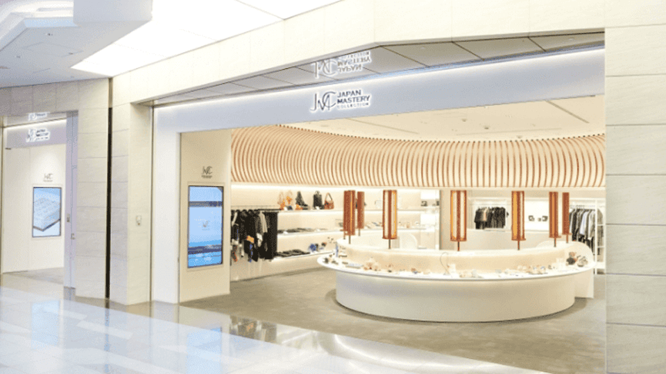 羽田空港国際線にある MIZEN ストアの明るく開放的な店内外観。白を基調とした空間に曲線的な木製パネルの天井装飾が映え、中央の丸いディスプレイ台や壁面ラックに衣類や工芸品が整然と並んでいる。洗練されたデザインが、上質な日本ブランドを扱う店舗イメージを伝えている。